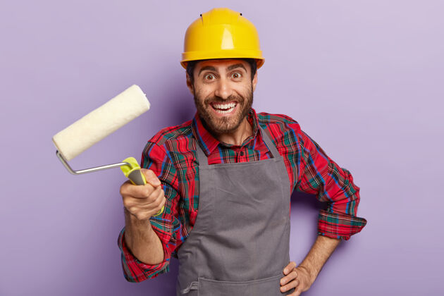 欢乐快乐迷人的工头准备修房子 拿着油漆辊 装饰墙壁 戴着黄色的防护帽 格子衬衫和围裙 积极地微笑着拿着建筑工具的男人积极的辛勤工作装饰
