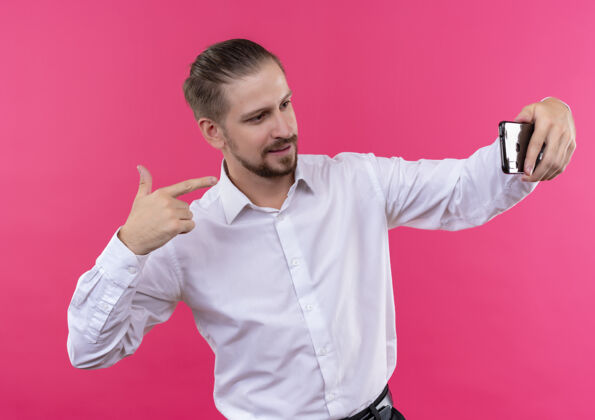 微笑穿着白衬衫的英俊商人站在粉色背景下微笑着用智能手机自拍手机自拍白色