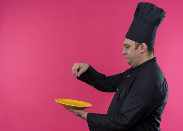 脸男厨师身穿黑色制服 头戴厨师帽 在盘子上撒盐 表情严肃 站在粉色背景上粉盘子男