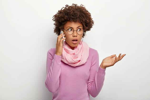 卷发困惑的非洲发型女性 与同事或朋友交谈 试图通过智能手机解决问题 注意力集中在一边 戴眼镜 紫色休闲套头衫 脖子上围着围巾肖像不高兴惊喜