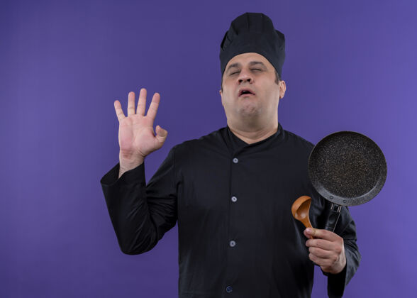 厨师穿着黑色制服 头戴厨师帽 闭着眼睛站在紫色背景上拿着煎锅的男厨师薯条烹饪立场