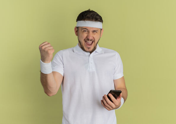 衬衫身穿白衬衫 头戴智能手机的年轻健身男子高举拳头站在橄榄色背景下 快乐而兴奋拳头白色举行