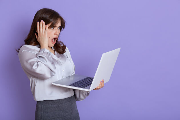 不快乐迷人的紧张的年轻女商人 手里拿着笔记本电脑 用震惊的表情看着设备的屏幕 用手抚摸着她的头 张大嘴巴 靠着淡紫色的墙壁站着计算机商业年轻