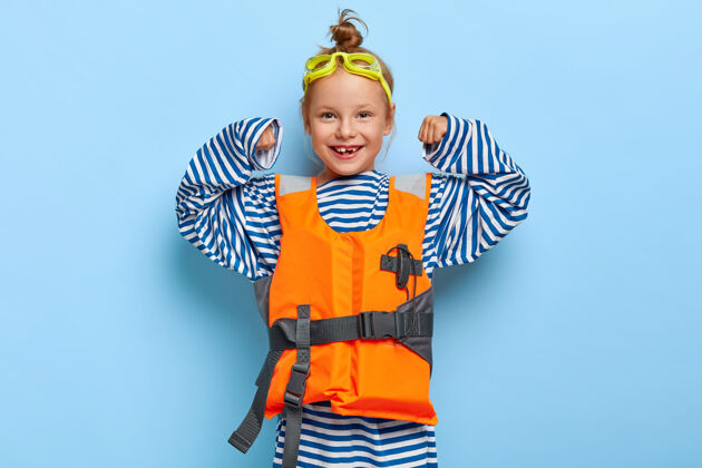 手臂穿着宽松条纹水手套头衫的可爱红发女孩 举起双臂 展示自己的力量 假装是海上救生员 戴着护目镜和充气救生衣 展示力量巡航站红发