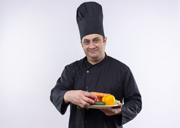 男男厨师身穿黑色制服 头戴厨师帽 手持盛有新鲜蔬菜的盘子 站在白色背景下 面带微笑地看着镜头盘子穿看