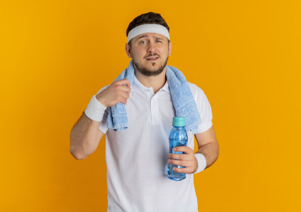 健身身穿白衬衫 头上系着头带 脖子上围着毛巾 手里拿着一瓶水 站在橙色背景下 自信地看着镜头站着拿着水