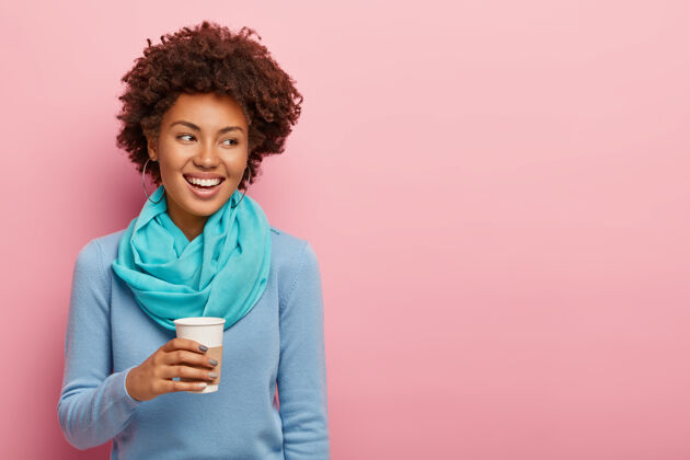 女性一个长着一头非洲头发的开朗可爱的女人喝着外卖咖啡 享受着辛苦工作后的休息 有着愉快的谈吐 带着牙齿般的微笑站在一旁 穿着蓝色的衣服 隔离在粉色的墙上休闲套头衫外卖