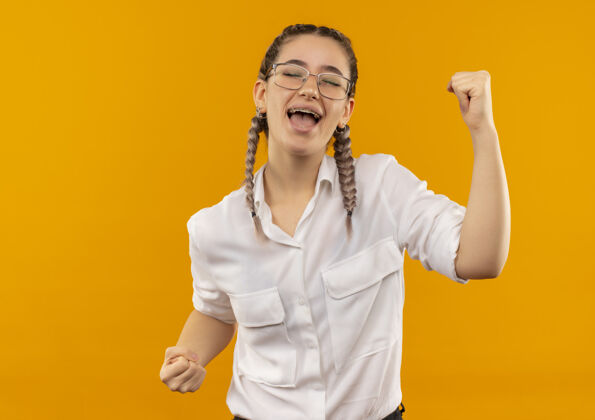 辫子戴着眼镜 梳着辫子 穿着白衬衫 握紧拳头的年轻女学生站在橙色的背景下为自己的成功而高兴和兴奋白色学生女孩
