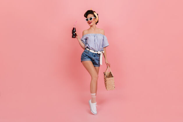 女士身材匀称的小姑娘手里拿着一瓶苏打水 面带微笑迷人的欧洲女人拿着一个夏天的袋子 在粉色的背景上摆姿势优雅玩苏打水
