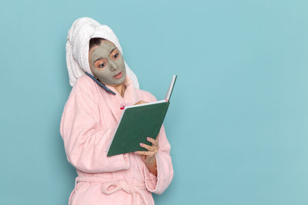 正面正面图身着粉色浴袍的年轻女性淋浴后讲电话阅读淡蓝色墙壁上的抄写本美丽的水自我护理淋浴干净浴袍笔记本电脑清洁
