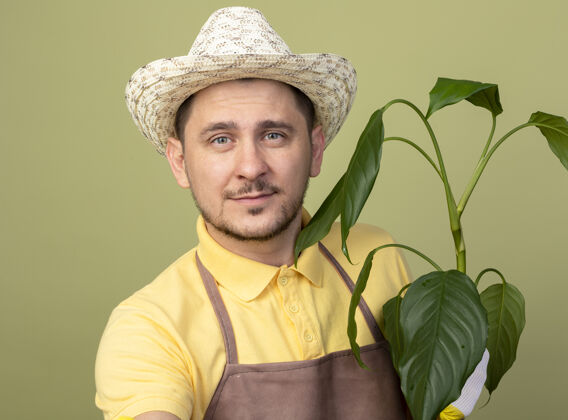 人年轻的园丁戴着工作手套 穿着连体衣 戴着帽子 拿着植物 面带微笑地看着前面 站在明亮的墙上制服穿脸