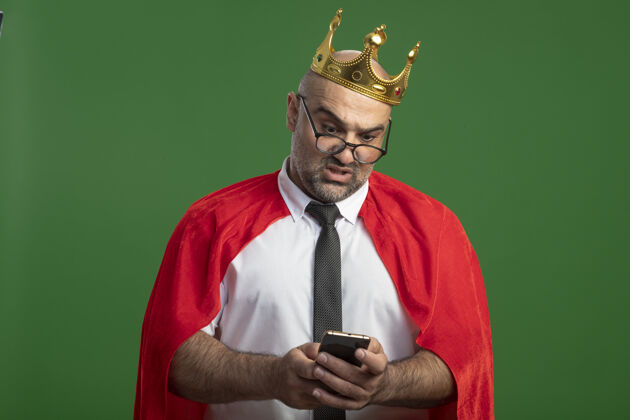 人穿着红色斗篷戴着皇冠的超级英雄商人站在绿色的墙上 用智能手机看着困惑商人手机面部表情