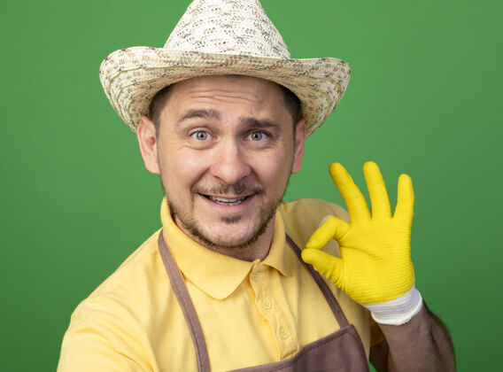 人年轻的园丁穿着连体衣 戴着帽子 戴着工作手套 站在绿色的墙边 看着前面的ok标志 开心地微笑着工人站人