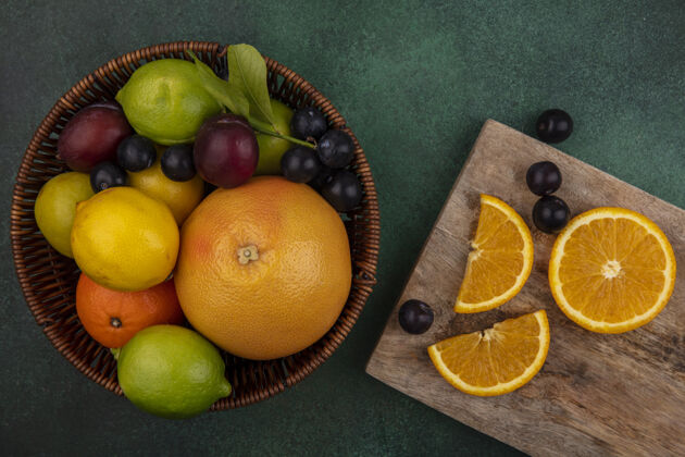李子俯视图在一个绿色背景的篮子里放着柚子 樱桃 李子 柠檬 酸橙和李子的砧板上的橘子片橘子葡萄柚切片