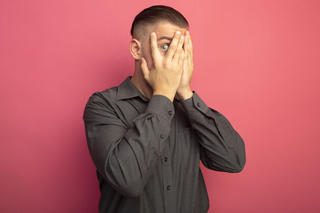 人一个穿着灰色衬衫的年轻帅哥站在粉红色的墙上 用手遮住眼睛 用手指看着姿势手衬衫