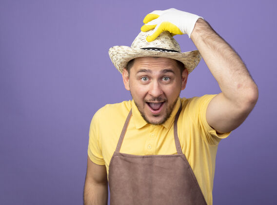 看年轻的园丁穿着连体衣 戴着工作手套戴着帽子 面带微笑 兴高采烈地抚摸着站在紫色墙上的帽子连身衣制服帽子