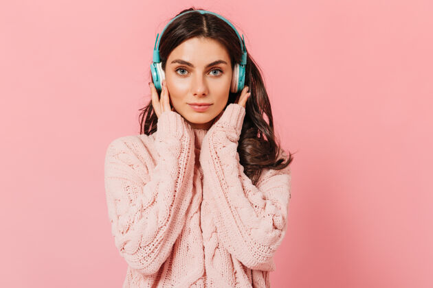 女性穿着针织毛衣的女孩按下耳机以获得更好的声音蓝眼睛的女人面带微笑地看着粉色背景的相机衣服啦啦队耳机