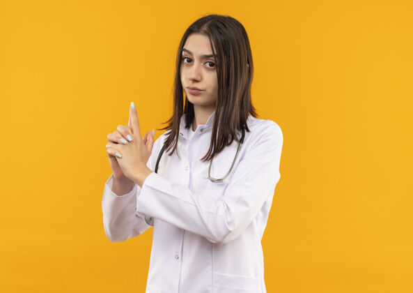 外套年轻的女医生穿着白大褂 脖子上戴着听诊器 用手指朝前做着或枪的手势 严肃的脸站在橙色的墙上医生医院年轻人