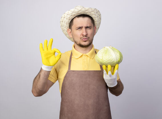 工作年轻的园丁 穿着连体衣 戴着帽子 戴着工作手套 手里拿着卷心菜 自信地看着前面 站在白墙上的ok标志连身衣展示制服