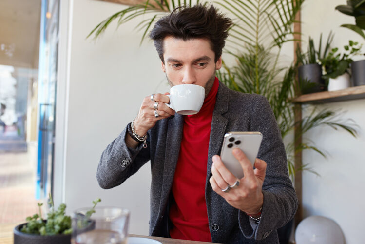 夹克一张室内照片 年轻漂亮的棕色头发的男人穿着优雅的衣服 一边在城市咖啡馆喝咖啡 一边举起手拿着智能手机 积极地看着屏幕午餐未剃毛衣服