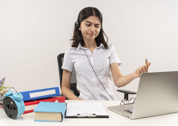 手势穿着白衬衫 戴着耳机的年轻女孩 坐在桌子旁 手里拿着文件夹和笔记本电脑 满脸不快坐着女孩衬衫