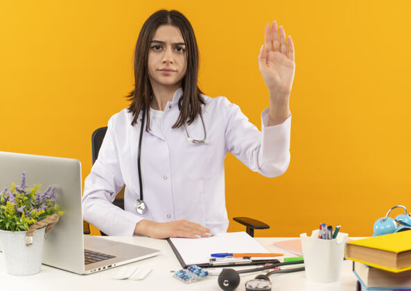 停止年轻的女医生穿着白大褂 手持听诊器 面带严肃的表情 手拿着笔记本电脑和文件坐在桌子旁 站在橙色的墙上 朝前面看坐着笔记本电脑外套