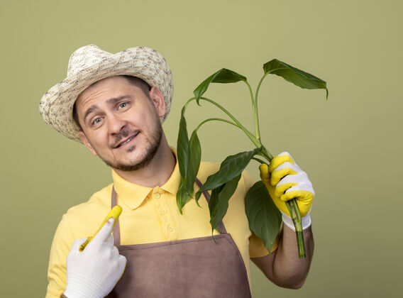 看年轻的园丁 穿着连体衣 戴着帽子 戴着工作手套 手里拿着一株植物 站在明亮的墙上 面带微笑地看着前方年轻人人快乐