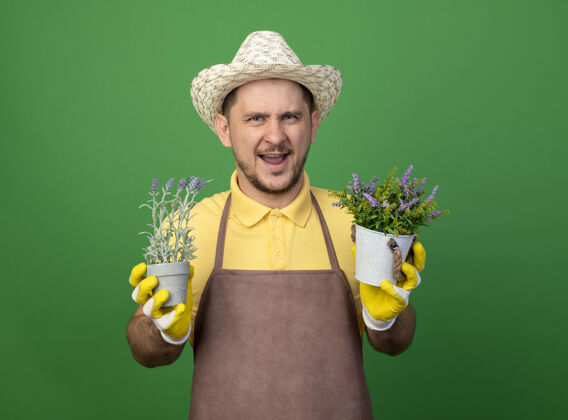 植物年轻的园丁穿着连体衣 戴着帽子 戴着工作手套 手里拿着盆栽植物 站在绿色的墙边愉快地微笑着看着前方年轻人花园手套