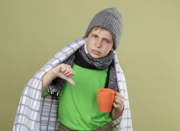 拇指生病的小男孩穿着绿色t恤 围着暖和的围巾 帽子裹在毯子里 手里拿着一杯热茶 大拇指朝下 站在明亮的墙上 不开心 生病围巾小茶