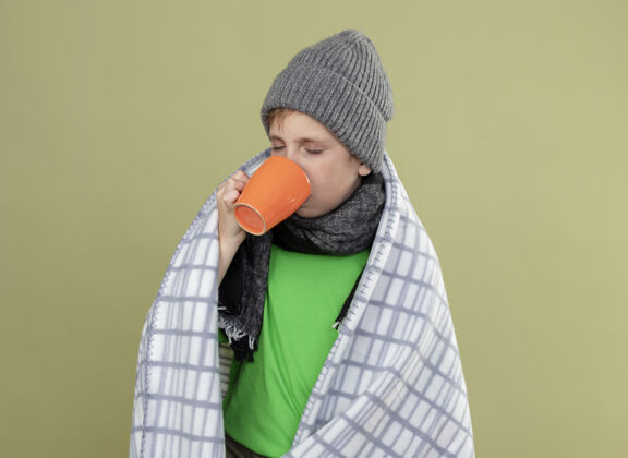 温暖生病的小男孩穿着绿色t恤 戴着暖和的围巾和帽子 裹着毯子 站在轻墙上喝着热茶包小毯子