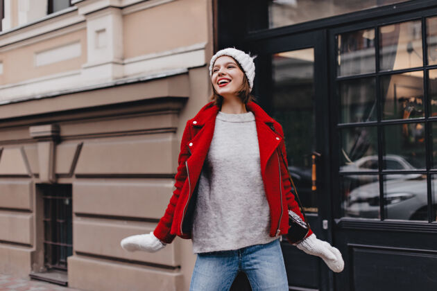 冷穿着牛仔裤 灰色毛衣 红色外套 戴着手套的针织帽子的漂亮女孩在街上摆着微笑的姿势时尚年轻地图