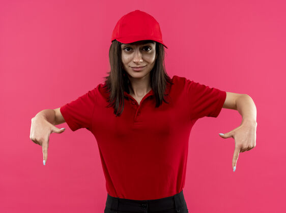 交货年轻的送货员穿着红色马球衫 戴着帽子 食指朝下 微笑着自信地站在粉红色的墙上穿立场马球