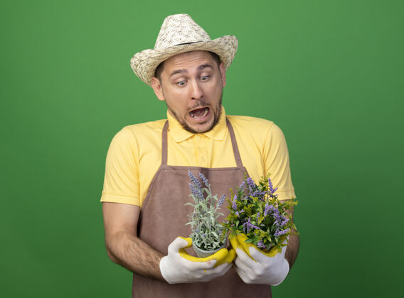 人年轻的园丁穿着连体衣 戴着帽子 戴着工作手套 手里拿着盆栽植物 站在绿色的墙壁上惊讶地看着它们人花园锅