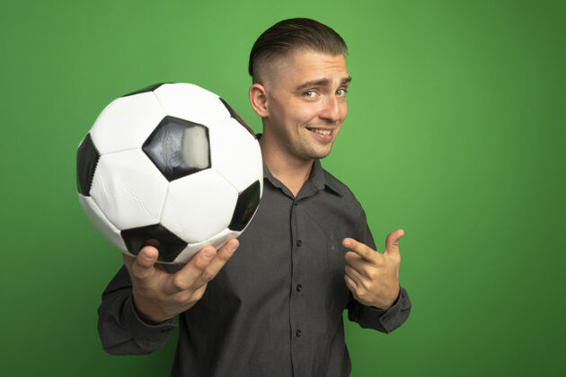 微笑身着灰色衬衫的年轻帅哥展示着足球 食指指着足球微笑着站在绿色的墙上手指指向衬衫