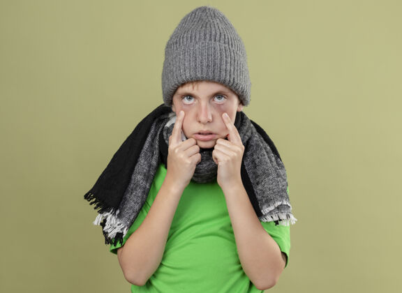 光生病的小男孩穿着绿色t恤 戴着暖和的围巾 戴着帽子 站在明亮的墙上手指着眼睛 看起来很不舒服看手指疾病