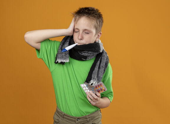 暖气生病的小男孩穿着绿色t恤 脖子上围着一条暖和的围巾 嘴里叼着体温计 站在橙色的墙上 手里拿着看起来不舒服的药片疾病小橘子
