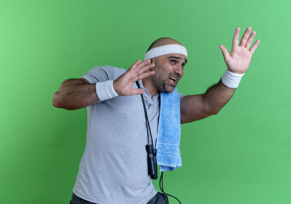 手戴着头巾 脖子上围着毛巾的成熟的运动型男人 带着恐惧的表情 站在绿色的墙上做着防御的手势人毛巾恐惧