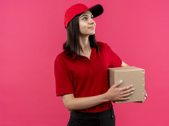 看年轻的送货员身穿红色马球衫 头戴鸭舌帽 手拿纸盒包装 面带微笑地站在粉红色的墙上 看着一旁马球站着放在一边
