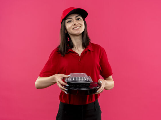 抱身穿红色马球衫 头戴帽子 手拿食物包的年轻送货女孩友好地站在粉红色的墙上帽子站女孩