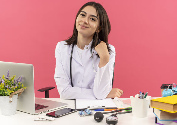 周围年轻的女医生穿着白大褂 脖子上戴着听诊器 面带微笑 自信地坐在桌旁 粉色的墙上挂着笔记本电脑人坐着办公室