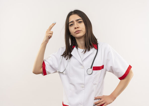 女人年轻的女医生穿着白大褂 脖子上戴着听诊器 不高兴地看着前面 两个手指站在白墙上外套年轻人手势