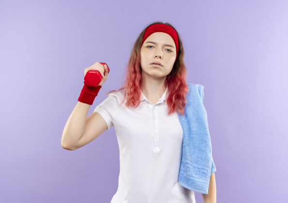 严肃年轻的运动型女人 肩上扛着毛巾 举着一个哑铃 严肃的脸站在紫色的墙上做练习市民抱着运动员
