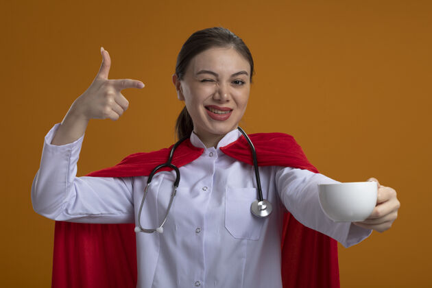 人类女医生手持听诊器 身穿白色医疗制服 身穿红色超级英雄斗篷 站在橙色的墙上 用手指指着咖啡杯 微笑着眨眼医疗杯子人