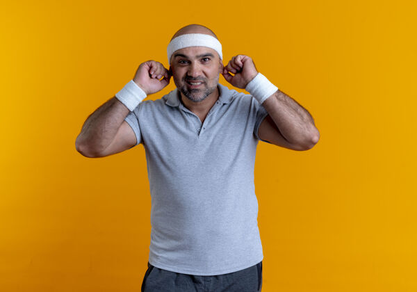 运动员戴着头巾的成熟的运动型男人微笑着看着前面 他的耳朵站在橙色的墙上微笑教练人