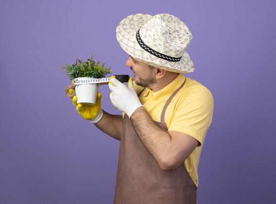 连身衣年轻的园丁穿着连体衣 戴着帽子 戴着工作手套 手里拿着盆栽植物 用卷尺量着它 站在紫色的墙上显得很好奇男性人人