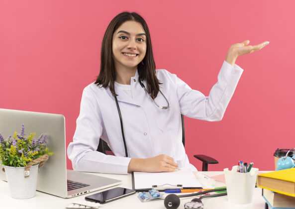 桌子年轻的女医生穿着白大褂 脖子上戴着听诊器 坐在桌旁用手提电脑对着粉色的墙壁微笑着展示着什么脖子胳膊外套