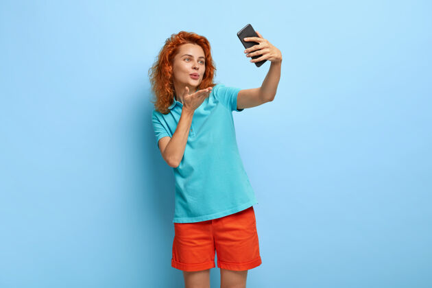 手机可爱的年轻女模特在手机摄像头前送上飞吻 为男友做自拍肖像分享照片GingerSwoosh科技