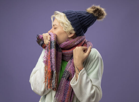 女人戴着暖和帽子和围巾的短发生病的不健康的女人在紫色的墙上咳嗽头发帽子温暖