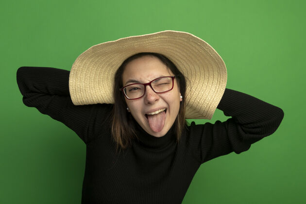 夏天年轻漂亮的女人戴着夏天的帽子 穿着黑色高领毛衣 戴着眼镜 看着前面伸出的舌头 快乐而积极地站在绿色的墙上看姿势人类