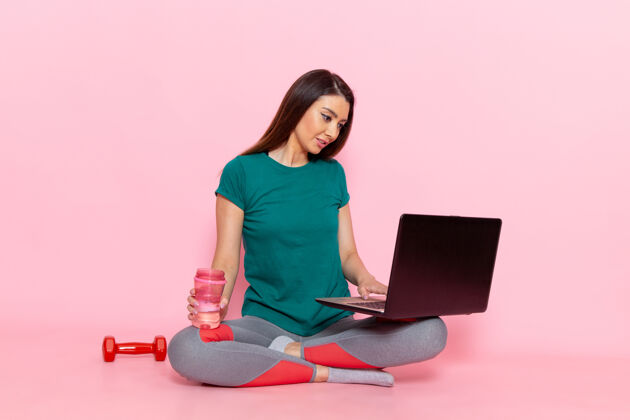 运动正面图身着绿色t恤的年轻女性在浅粉色墙壁上使用笔记本电脑腰部运动锻炼美丽苗条的运动员女性女性运动便携式电脑